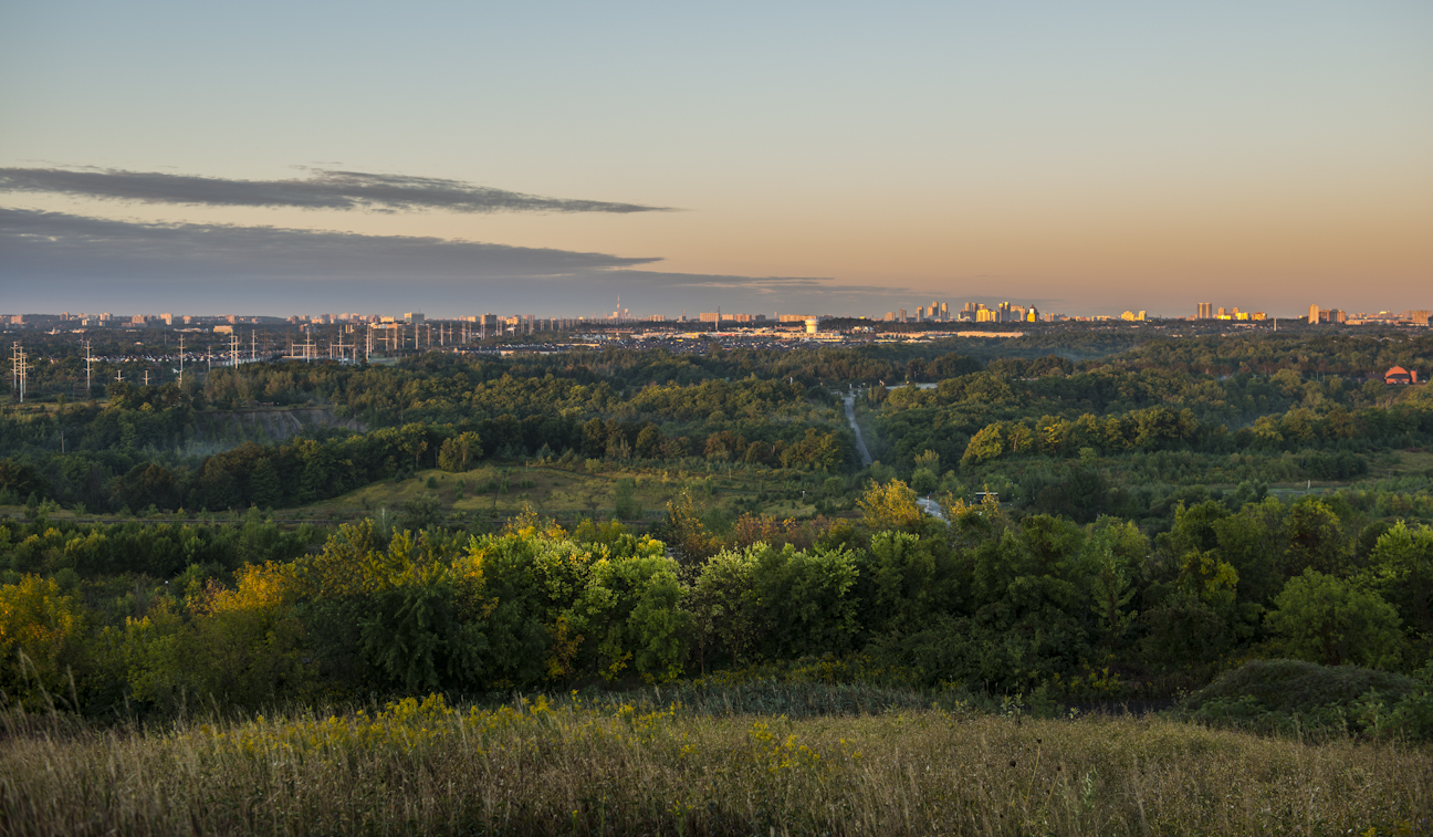 Cette photographie montre les divers paysages du parc urbain national de la Rouge et des alentours, qui passent de milieux naturels non perturbés à des paysages aménagés et urbains en arrière-plan. En avant-plan, on voit un pré résiduel, qui laisse progressivement place à une zone de ravins densément boisée. À l’horizon se dresse la métropole de la région du Grand Toronto, avec ses nombreux gratte-ciel et la tour du CN. Une grange rouge est visible à droite de la photo, alors qu’au centre, une longue route sinueuse traverse le ravin boisé.